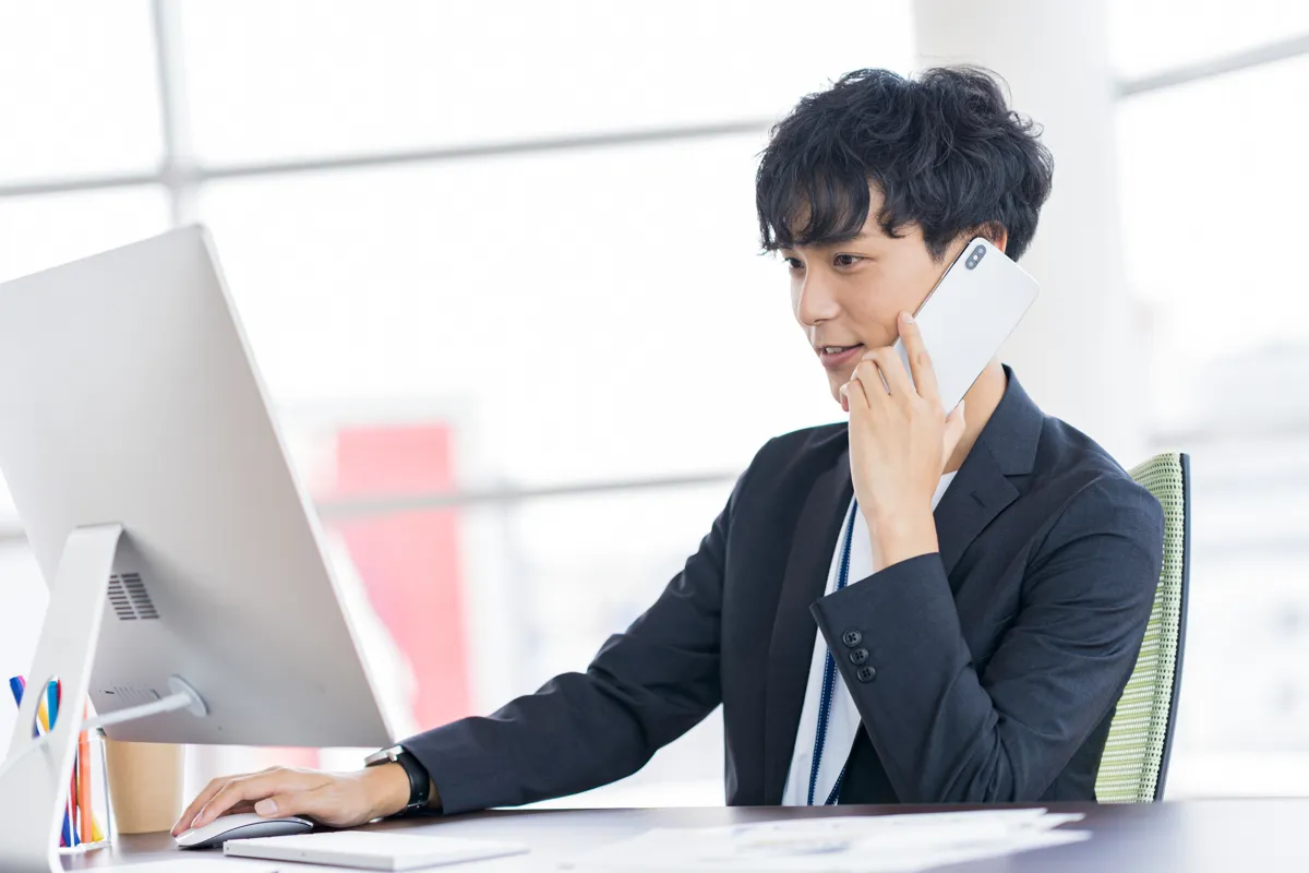 スーツ姿の男性が電話を受けながらパソコンを操作している様子。